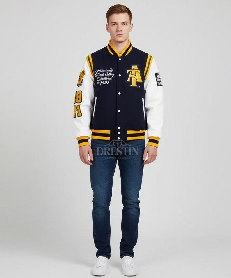 May include: Navy blue and white varsity jacket with yellow accents. Features the letter "A" and text on the chest and sleeve. The jacket has a button-up front and ribbed cuffs. The model is wearing blue jeans and white sneakers.