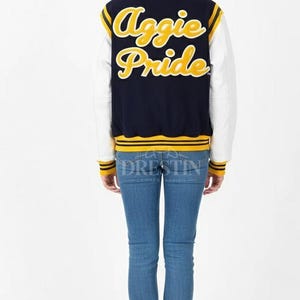 May include: A navy blue and white letterman jacket with the words "Aggie Pride" in yellow script on the back. The jacket has yellow and white striped accents and is worn with blue jeans and white sneakers.
