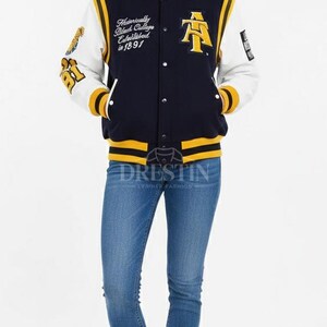 May include: A navy blue and white varsity jacket with yellow accents. The jacket features text on the chest and sleeve, and is paired with blue jeans and white sneakers. The jacket has a button-up front.