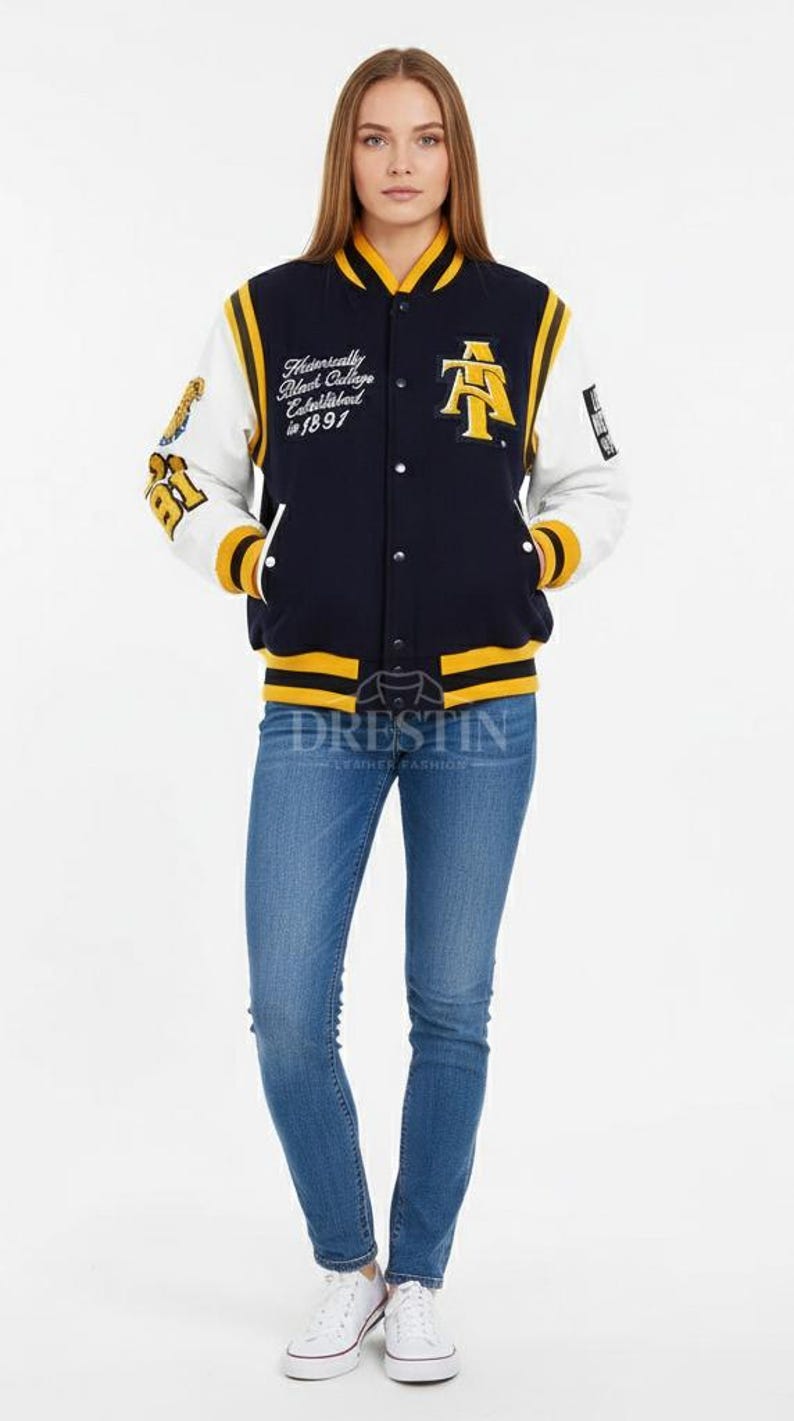 May include: A navy blue and white varsity jacket with yellow accents. The jacket features text on the chest and sleeve, and is paired with blue jeans and white sneakers. The jacket has a button-up front.