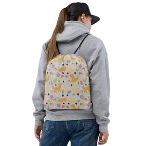 May include: A light gray drawstring backpack featuring a repeating pattern of golden retriever dogs, paw prints, mushrooms, and flowers. The bag has black drawstrings and is worn over the shoulders.