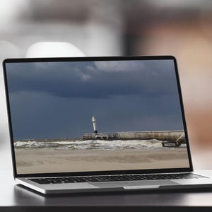 Puede incluir: Un portátil plateado muestra una escena costera con un faro, un muelle y un cielo tormentoso. El portátil está abierto sobre una superficie oscura, con un fondo borroso y una silla blanca visible.