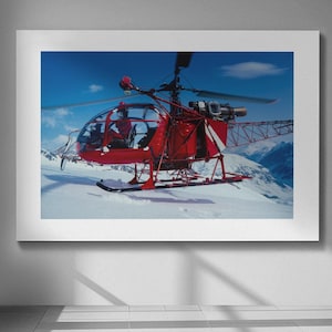 May include: A framed image of a red helicopter on a snowy mountain. The helicopter has a clear cockpit and is set against a blue sky with clouds. The image is displayed on a white wall in a room with large windows.