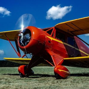 May include: A classic red and yellow biplane with a large propeller, parked on a grassy field under a blue sky with fluffy white clouds. The plane has a vintage design with a rounded engine cowling and yellow wings.