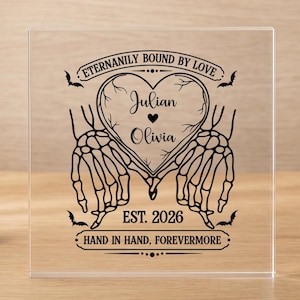Op de afbeelding: Een vierkante, transparante acrylplaat met een zwarte opdruk van twee skelettenhanden die een hartvorm maken. De tekst luidt ETERNALLY BOUND BY LOVE, Julian, Olivia, EST. 2026 en HAND IN HAND, FOREVERMORE, versierd met kleine vleermuisjes.