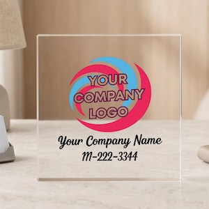 May include: Clear acrylic block with a company logo in pink and blue. The text "YOUR COMPANY LOGO" is above "Your Company Name" and a phone number. The block is on a desk.