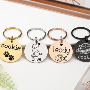 May include: Four round pet ID tags with key rings. The tags are gold, silver, rose gold, and black. Each tag has a different design: paw prints, a dog silhouette, a fish, and a planet with the name "cookie" on two of the tags.
