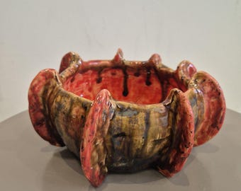 Puede incluir: Un cuenco de cerámica hecho a mano con un diseño único y orgánico. El cuenco presenta un interior rojo vibrante con goteos oscuros, y el exterior muestra una mezcla de tonos marrones, amarillos y rojos. El cuenco tiene una superficie texturizada y es adecuado para uso decorativo.
