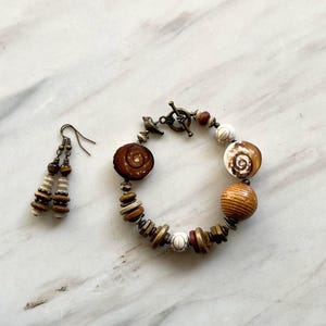 Puede incluir: Un conjunto de joyas hechas a mano que incluye una pulsera y pendientes a juego. La pulsera tiene un cierre de palanca y está hecha con cuentas de madera, cuentas en forma de concha y detalles en marrón, blanco y beige.