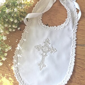 May include: White baby bib with a decorative cross and lace trim. The bib features a satin finish and a delicate embroidered cross design. A satin ribbon is used to tie the bib around the neck. The bib is trimmed with a delicate lace.