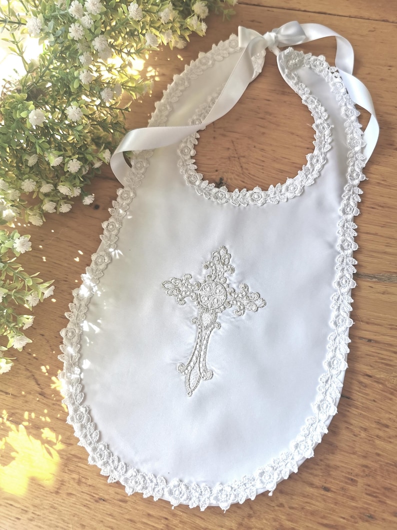 May include: White baby bib with a decorative cross and lace trim. The bib features a satin finish and a delicate embroidered cross design. A satin ribbon is used to tie the bib around the neck. The bib is trimmed with a delicate lace.