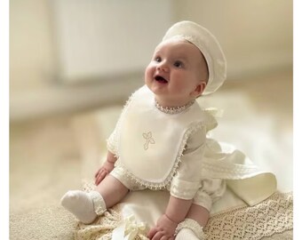 Peut inclure: Une robe de baptême blanche pour bébé avec une bordure en dentelle et une croix brodée sur la bavette. Le bébé porte un chapeau blanc et des chaussettes blanches.