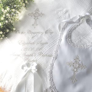 May include: A white baptism set featuring a blanket, bib, and ribbon. The blanket and bib are adorned with embroidered crosses and lace trim. The blanket has text that reads "On the Confirmation Day of Elizabeth Paige".