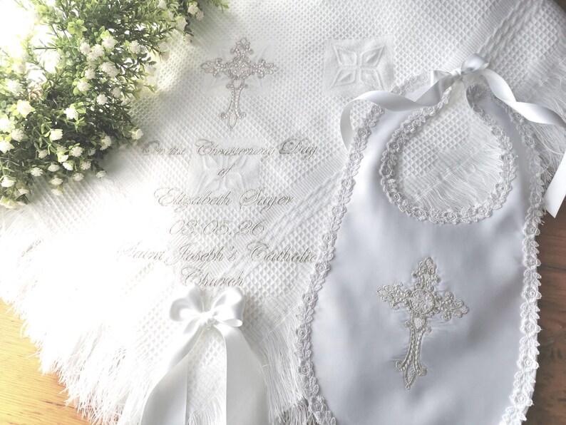 May include: A white baptism set featuring a blanket, bib, and ribbon. The blanket and bib are adorned with embroidered crosses and lace trim. The blanket has text that reads "On the Confirmation Day of Elizabeth Paige".