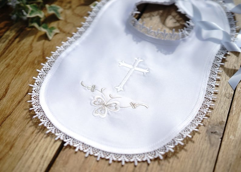 May include: White baby bib with a delicate lace trim and a cross and floral embroidery. The bib has a satin ribbon for tying. A beautiful baptism or christening bib.