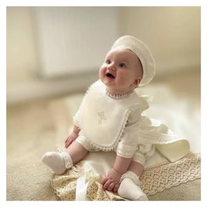 May include: A baby wearing a cream-colored christening outfit, including a bib with a cross, a beret, and matching socks. The outfit is trimmed with lace and the baby is sitting on a lace-edged blanket.