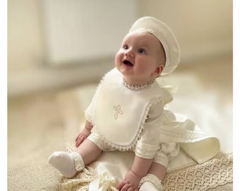 Peut inclure: Une robe de baptême blanche pour bébé avec une bordure en dentelle et une croix brodée sur la bavette. Le bébé porte un chapeau blanc et des chaussettes blanches.