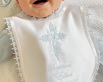 Peut inclure: Bavette de baptême en satin blanc avec une croix brodée bleue et le texte "Baptême d'Elliot 12 mars 2022". La bavette est bordée de dentelle.