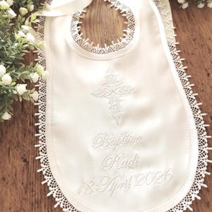 May include: White satin baby bib with a delicate lace trim and a cross embroidered in the center. The bib features the embroidered text "Baptism Radi 18 April 2026" and a satin ribbon tie.