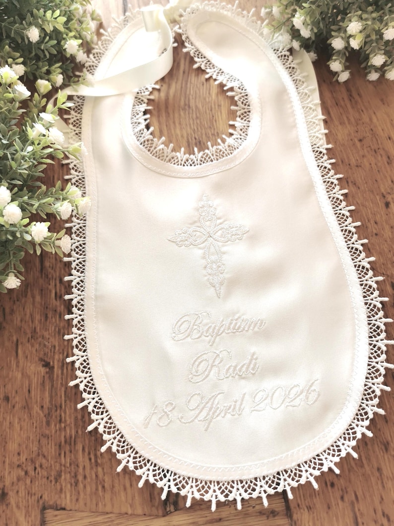 May include: White satin baby bib with a delicate lace trim and a cross embroidered in the center. The bib features the embroidered text "Baptism Radi 18 April 2026" and a satin ribbon tie.