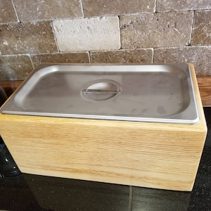 May include: A wooden box with a stainless steel insert. The box is light brown and has a wood grain pattern. The insert has a rounded edge and a handle.
