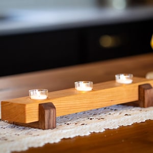 May include: A wooden candle holder with three tealight candle holders. The holder is made of light brown wood with dark brown accents. The candles are lit and glowing.
