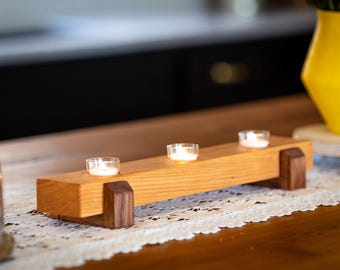 Pode incluir: Um porta-velas de madeira com três porta-velas para velas pequenas. O porta-velas é feito de madeira marrom claro com detalhes marrons escuros. As velas estão acesas e brilhando.