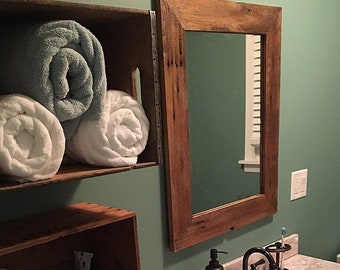 May include: A rustic wooden framed mirror with a light wood finish hangs above a bathroom sink.  The mirror is mounted on a green wall.  Towels are rolled and stacked on a wooden shelf.