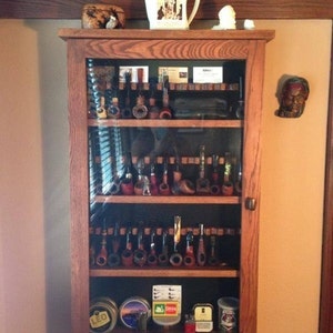May include: A wooden cabinet with glass doors displays a collection of pipes. The cabinet has three shelves and is filled with various pipes, some with decorative details. The cabinet is made of dark wood and has a simple design.