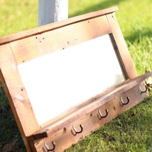 May include: A rustic wooden mirror with a shelf and hooks. The mirror is rectangular and has a distressed wood frame. The shelf is made of reclaimed wood and has a natural finish. The hooks are made of metal and are attached to the bottom of the shelf. The mirror is leaning against a white fence in a grassy area.