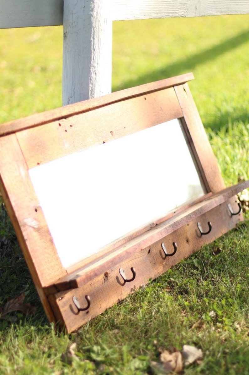 May include: A rustic wooden mirror with a shelf and hooks. The mirror is rectangular and has a distressed wood frame. The shelf is made of reclaimed wood and has a natural finish. The hooks are made of metal and are attached to the bottom of the shelf. The mirror is leaning against a white fence in a grassy area.