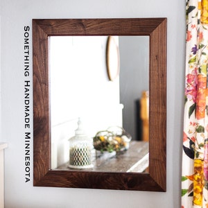 May include: A rectangular mirror with a dark brown wooden frame. The frame is thick and has a simple design. The mirror is hanging on a white wall. The text "SOMETHING HANDMADE MINNESOTA" is visible on the wall to the left of the mirror.
