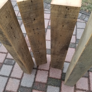 May include: Four reclaimed wood beams, each with a different grain pattern and color variation. The beams are weathered and have a rustic appearance. They are arranged in a fan shape on a gray brick surface.