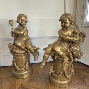 Könnte beinhalten: Zwei antike, goldfarbene Cherubinstatuen. Einer der Cherubim hält einen Pfeil, der andere einen Bogen. Beide sitzen auf verzierten Sockeln mit Girlandendetails. Die Statuen sind aus Metall und werden in Innenräumen ausgestellt.