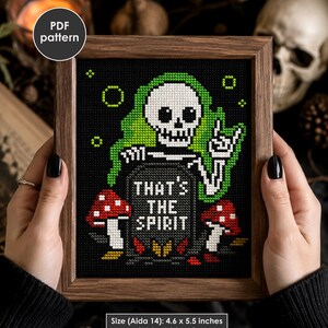 May include: A cross-stitch pattern in a wooden frame featuring a white skeleton with green hair making a rock-and-roll hand gesture behind a tombstone that reads THAT'S THE SPIRIT, flanked by two red and white mushrooms. Size is 4.6 x 5.5 inches.