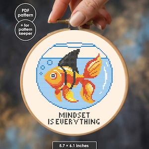 May include: A cross-stitch pattern in a wooden hoop featuring a goldfish wearing a black shark fin inside a blue fishbowl. The text reads "MINDSET IS EVERYTHING". A label indicates dimensions of 5.7 x 6.1 inches and includes PDF pattern details.