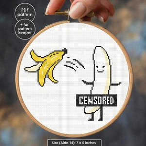 以下が含まれることがあります： 木製の刺繍枠に入ったクロスステッチパターン。黄色いバナナの皮と、CENSOREDと書かれた黒い帯を身につけた白いバナナのキャラクターが描かれています。デザインにはPDF patternとpattern keeperの文字が含まれ、サイズは17 x 13 cmです。