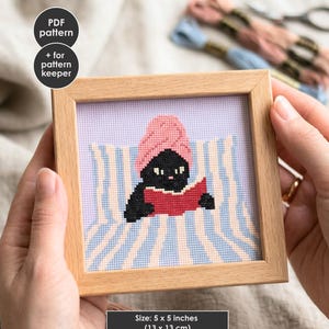 May include: A cross-stitch pattern in a light wood frame featuring a black cat with a pink towel on its head reading a red book while lying on a blue and white striped towel. The text reads PDF pattern + for pattern keeper. Size: 5 x 5 inches.