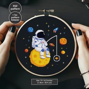 May include: A cross-stitch embroidery hoop featuring an astronaut fishing on a yellow planet against a black background with stars and planets. The design includes a rocket ship and the text "PDF pattern + for pattern keeper". The size is 7.9 x 8.2 inches.