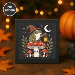 May include: A framed cross-stitch pattern featuring a green frog wearing a witch hat, sitting on a red mushroom with white spots. The frog holds a lantern under a crescent moon and stars. The words "PDF pattern" are in a white circle.