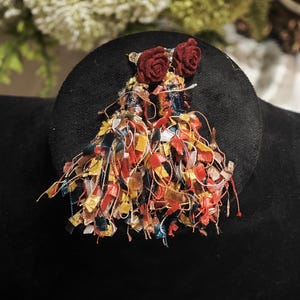 May include: A pair of handmade earrings featuring small red rose accents and colorful tassel fringe. The earrings are attached to a black circular base, creating a striking contrast. The tassels incorporate various colors, including yellow, blue, and red.
