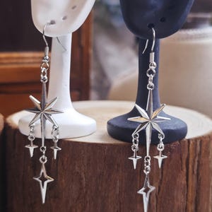 May include: Silver-toned dangle earrings featuring a starburst design. Each earring has a large star at the top with smaller stars dangling below. The earrings are displayed on white and black earring stands.