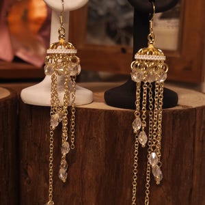 May include: Gold-toned chandelier earrings with a crown-like top, adorned with small white beads and dangling chains. Clear, teardrop-shaped crystals are interspersed along the chains. The earrings are displayed on white and black earring stands.