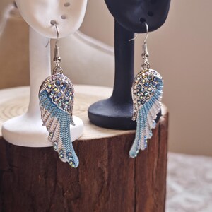 May include: A pair of silver-toned earrings in the shape of angel wings. The earrings feature a blue and white enamel design with iridescent rhinestones. The wings are displayed on white and black earring display stands.