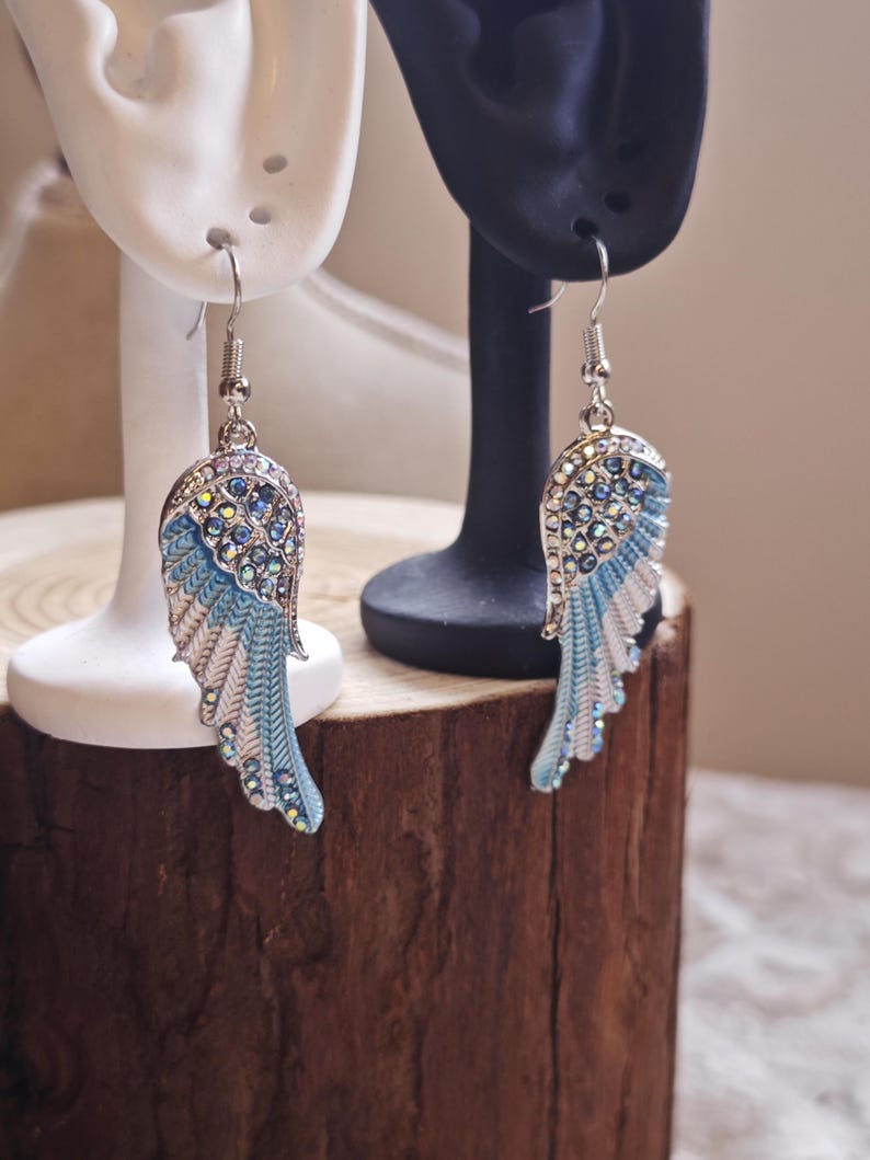 May include: A pair of silver-toned earrings in the shape of angel wings. The earrings feature a blue and white enamel design with iridescent rhinestones. The wings are displayed on white and black earring display stands.