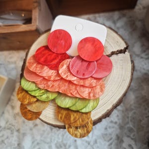 May include: Colorful, layered disc earrings in shades of red, orange, green, and gold. The earrings are displayed on a white backing and a wooden slice, showcasing their vibrant, eye-catching design. These statement earrings are perfect for adding a pop of color.