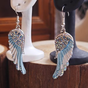 May include: A pair of silver-toned wing earrings with blue and white enamel and sparkling crystal accents. The earrings are displayed on white and black earring stands, set against a wooden background.
