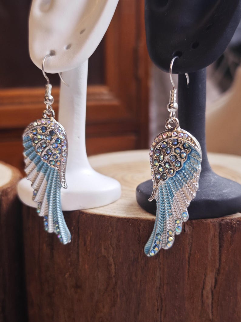 May include: A pair of silver-toned wing earrings with blue and white enamel and sparkling crystal accents. The earrings are displayed on white and black earring stands, set against a wooden background.