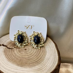 Può includere: Un paio di orecchini a lobo color oro con una pietra ovale scura al centro. Gli orecchini hanno un design decorativo a forma di foglia attorno alla pietra. Gli orecchini sono esposti su una carta bianca con le lettere "SZJ".