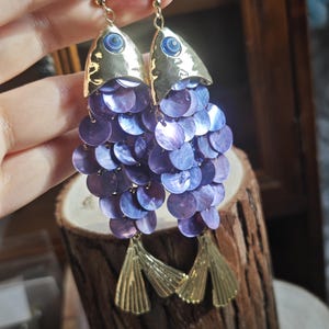 Può includere: Un paio di orecchini a forma di pesce. Gli orecchini presentano teste di pesce in metallo color oro con dettagli occhi blu. I corpi sono fatti di paillettes viola iridescenti e pinne in metallo color oro pendono sotto.